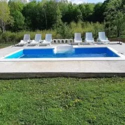 Rechteckiger Pool mit vier weißen Sonnenliegen neben einer Rasenfläche und einem Wald